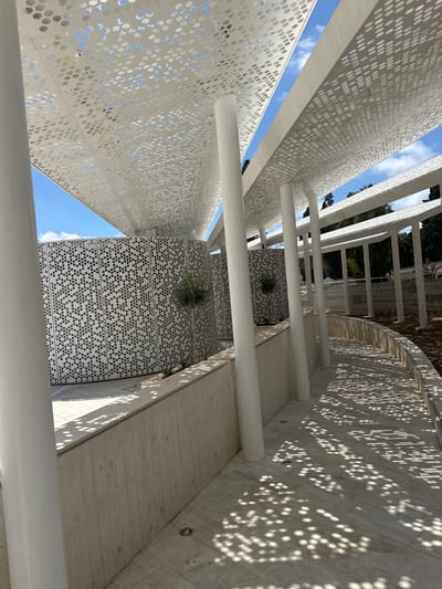The image depicts an outdoor walkway with a curved retaining wall on the right and a patterned, perforated white canopy overhead. The canopy casts intricate shadows on the walkway. On the left, there is a curved wall with a geometric, honeycomb-like pattern of perforations. A small, young olive tree is planted in front of this patterned wall. The walkway is paved with light-colored square tiles. The background shows trees and a cloudy blue sky. This architectural feature is part of the Kato Paphos Archaeological Park in Paphos Municipality, Cyprus. There are no people visible in the image.