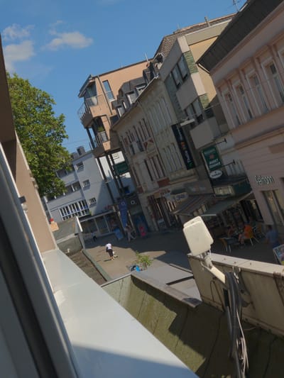 The image shows a street scene in Kamen, Germany. The perspective is from an elevated position, looking down at an angle. In the foreground, there is a white structure, possibly part of a building or an outdoor fixture, with visible cables attached. Below and to the right of this structure, a street is visible with several people walking and sitting at outdoor tables.
Buildings line the street, displaying a variety of architectural styles and signage. Some of the visible signs are in German, including "Thalia" and "Markt Schanke." One building features a balcony with railings. Trees, a large green one on the left and a smaller one on the right, are present, as is a blue sky with some clouds. The overall impression is of a sunny day in a European town.