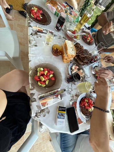 The image depicts an outdoor gathering at a table, possibly in Ravadinovo, Bulgaria, given the context provided. A table laden with food and drinks is the central focus. On the table, there is a large piece of grilled meat, sliced bread, a salad of tomatoes and cucumbers, a dish with what appears to be cheese and some red seasoning, nuts in a bowl, and various glasses filled with drinks, including what looks like lemonade and dark liquid. A bottle of olive oil stands upright. Two hands are visible actively pouring drinks into glasses, suggesting an ongoing toast or serving. In the foreground, a person wearing blue jeans and a white t-shirt is partially visible, with a mobile phone and a pack of Karelia cigarettes on the table nearby. The table is covered with a floral-patterned tablecloth. The background shows greenery and hints of an outdoor setting. The overall scene conveys a casual meal and social interaction.