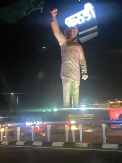 A nighttime photograph captures a large, illuminated statue of a man in traditional Indian attire, with his right fist raised. The statue appears to be made of a reflective material, possibly metal or plastic, and is lit from below with green and white lights, casting a glow that illuminates the surrounding dark environment. Above the statue, a bright neon sign in Gurmukhi script glows white against the dark sky.

The statue stands on a platform, which is situated on what appears to be a highway median or a large vehicle. The base of the platform has more signage, also in Gurmukhi script, which is illuminated in yellow. The platform itself is bordered by a white and black striped barrier. In the background, streetlights and other distant lights suggest an urban or semi-urban setting at night. The overall impression is one of a monument or memorial, possibly commemorating a significant figure.

The location is identified as Jhiurheri, India. The photograph is taken from a slightly low angle, looking up at the imposing statue. The quality of the image is somewhat blurry, which is likely due to low light conditions and possibly movement during capture.