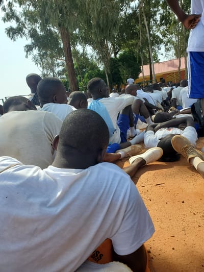 In Barakin Rafin Gora, Nigeria, a group of young people, predominantly boys and young men, are seated on the ground under trees. Many are wearing white t-shirts and some are in blue shorts. The ground is covered in a reddish-brown dirt. In the background, there is a building with an orange roof and a fence. The overall scene suggests an outdoor gathering or activity, possibly related to a school or training program.
