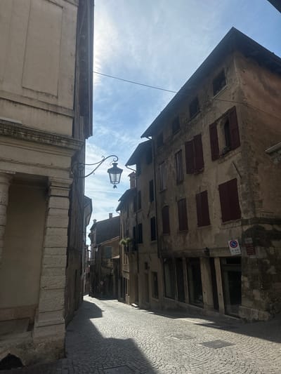 This is a vertical, eye-level, full shot of a cobblestone street in Maser, Italy, under a partly cloudy sky. The left side of the image is dominated by the corner of a light-colored stone building with large, arched openings. The right side features an aged, multi-story building with stucco walls, dark wooden shutters, and arched windows. A wrought-iron street lamp hangs from a bracket attached to the stone building on the left, extending over the street. The street is paved with small, gray cobblestones that reflect the sunlight. Shadows from the buildings stretch across the road. In the distance, the street curves and narrows, lined with more buildings. A street sign is visible on the right-hand building, with Italian text and a parking symbol. The overall atmosphere is one of historic European architecture bathed in sunlight.