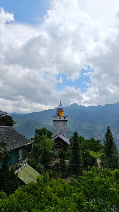 The image depicts a scene in Tả Van, Vietnam, featuring a stone and yellow lighthouse-like structure surrounded by lush green trees and vegetation. In the background, a range of mountains is visible under a cloudy sky with patches of blue. To the left, a portion of a green house with a dark gray roof is visible, partially obscured by the foliage. Another building with a gray roof is partially visible near the lighthouse. The scene is bathed in natural light, suggesting it is daytime.