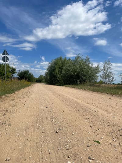 An eye-level, outdoor, medium shot captures a dirt road extending into the distance under a bright blue sky with scattered white clouds. The road, composed of light brown dirt and small stones, curves slightly to the left as it recedes. On the left side of the road, a tall, dark gray traffic sign with a white triangular border and a white circle inside is mounted on a pole, accompanied by electrical wires. Next to the sign, there are tall green grasses and yellow wildflowers. Further down the left side, a dense line of green trees and bushes is visible. On the right side of the road, a thick cluster of large, leafy green trees dominates the mid-ground, with a more slender, sparse tree to its right. Beyond the trees, a field with dry, light brown grass can be seen. The overall environment suggests a rural or semi-rural setting in Ракітніцкі сельскі Савет, Belarus. There are no people or animals visible in the image, and no actions or interactions are depicted.