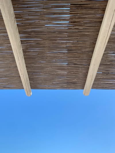 An upward view reveals the underside of a horizontally woven reed or bamboo shade structure, exhibiting brown tones. Two light-colored, natural wood support beams are visible, angling inward from the upper left and upper right corners towards the frame's center. Small gaps in the weaving allow slivers of bright sunlight to pass through. The lower half of the frame is occupied by a clear, bright blue sky. No individuals, additional objects, or discernible activities are present. The scene is an outdoor, sun-sheltered area in Φάρος, Greece.