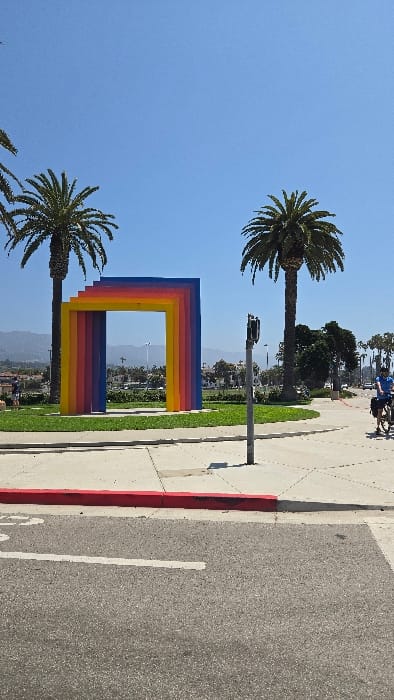 A vibrant, multi-hued rectangular art installation stands between two palm trees under a clear blue sky. The artwork is composed of stacked, progressively smaller frames. A street and a sidewalk edge the right side of the image, with people on bicycles visible in the distance. Hills can be seen through the opening in the art installation.