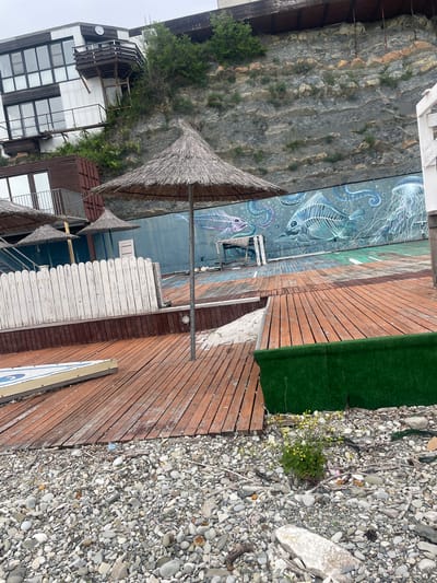 This image depicts a waterfront scene in Ol'ginka, Russia. A structure built on a cliff face dominates the left side of the image, with a mix of white and brown architecture. Several thatched umbrellas provide shade. The scene transitions to a wooden deck, which leads to an area covered in small, smooth stones, suggestive of a beach. Along the wall beside the deck, there is artwork featuring sea life. Some patches of artificial turf are scattered on the deck.