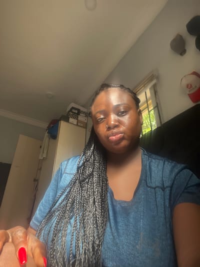 A person is lying on a dark surface. They are wearing a blue shirt and have braids. One of their hands is visible with red nail polish on the fingers. The location appears to be indoors, with a window visible and some objects on a shelf in the background. The city of Dushepe, Nigeria is not evident in the photo.