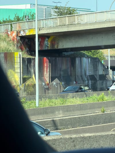 The image captures an urban scene with a focus on the underbelly of an elevated structure, likely a highway or bridge, in Rungis, France. The concrete supports of this elevated roadway are a dominant feature, with their weathered surfaces adorned with vibrant graffiti. The graffiti displays a mix of abstract patterns and stylized figures, primarily in bold colors like red, yellow, and black, creating a stark contrast with the duller concrete.
Below the elevated structure, a blue car is partially visible, suggesting that this area is accessible by road. Greenery, including bushes and grass, is growing along the edges of the concrete structures, indicating that nature is reclaiming parts of this man-made environment. The lighting suggests it is daytime, with natural light illuminating the scene. The overall mood is one of urban decay and artistic expression, where the raw structure of infrastructure is transformed by street art.
The image is oriented vertically, with the top of the frame showing a clear sky. Along the left side of the image, there are glimpses of metal structures with horizontal slats, possibly part of the elevated roadway's construction or adjacent buildings. The rough texture of the concrete and the rough application of paint in the graffiti are notable details. No people are directly visible in the image. The scene appears to be located in an outdoor, possibly semi-industrial or transit-oriented area given the presence of the highway structure.