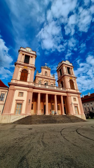 The image shows the Melk Abbey in Furth bei Göttweig, Austria, a magnificent Benedictine monastery. The abbey is a grand Baroque structure with two prominent bell towers, a central facade adorned with statues and a cross, and a portico with large columns. The building is painted in a pale pink and beige color scheme, with contrasting wooden doors and windows. A wide set of stairs leads up to the entrance.
In front of the abbey is a large, paved courtyard. The sky above is a vibrant blue, dotted with fluffy white clouds. The sun is shining, casting shadows and illuminating the architecture.
On the steps of the abbey, two individuals are seated, appearing to be resting or observing the surroundings. The ground in the courtyard shows some tire marks, suggesting recent activity, possibly from vehicles. A lamppost is visible on the left side of the building's base. The overall impression is one of historical grandeur and a peaceful, sunny day.