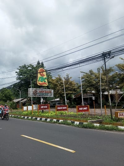 A road view shows a restaurant, "Ikan Bakar Cianjur," marked by a tall sign. Smaller signs in front advertise "Sop Nila," "Gurame Bakar," and "Nasi Liwet." A motorcycle with two riders passes on the road, marked by a yellow dividing line. Overhanging wires and a cloudy sky are visible above trees and the restaurant's structures.