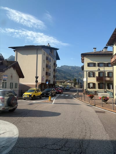 The image depicts a street scene in the town of Predazzo, Italy, likely during the daytime given the clear blue sky with some wispy clouds and the strong sunlight casting shadows. The perspective is from the street level, looking down a paved road that curves to the right.
On the left side of the street, a multi-story building with a light-colored facade and dark-tiled roof stands prominently. This building has balconies with railings and numerous windows. Adjacent to it is a smaller, older building with a darker roof and a decorative facade. Signs are visible on this smaller building, one with arrows indicating directions.
On the right side of the street, another multi-story building with a yellow facade and a dark-tiled roof is visible. This building also features balconies and shutters on its windows. Flower planters with colorful flowers are positioned along the sidewalk in front of this building.
Several cars are present on the road. A silver SUV is parked on the left. A bright yellow small car is stopped at what appears to be a pedestrian crossing or intersection in the middle of the road, along with a black SUV and a red car further down the road. Traffic lights and street lamps are also visible.
In the background, a mountainous landscape with forested slopes and rocky peaks dominates the horizon under the bright sky. The overall impression is of a peaceful, sun-drenched alpine town.