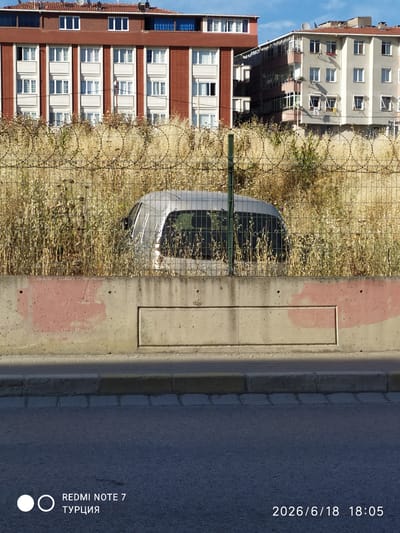 In Avcılar, Turkey, a silver vehicle is partially visible behind a barbed wire fence and overgrown dry grass. The vehicle appears to be abandoned or stored within a secured area. In the background, two multi-story buildings, one with a red-brick facade and the other with a light-colored exterior, dominate the cityscape. A concrete wall with pink paint marks runs across the lower portion of the image. The scene is captured during daylight, indicated by the clear sky and shadows. The timestamp "2026/6/18 18:05" is present in the lower right corner of the image, along with the text "REDMI NOTE 7 ТУРЦИЯ".