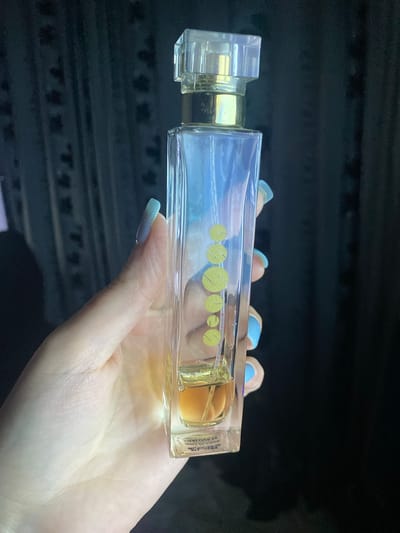 A person's hand, adorned with light blue and white ombre nail polish, holds a rectangular glass bottle horizontally against a dark, textured background resembling a car interior. The bottle, likely a perfume or cologne, features a clear, angular cap and a golden sprayer mechanism. The liquid inside is a light amber color. On the bottle's surface, there are five prominent yellow-green circular designs, arranged in a row, with a textured pattern within each circle.

Visible text on the bottle includes "PARFUM" and possibly "EAU DE PARFUM" near the sprayer, seen through the glass. On a label on the right side of the bottle, partial text can be deciphered as "30 ML e", "VOYAGE", and "DE PARFUM". Other smaller text on the label is illegible due to reflection and resolution.

The scene takes place indoors, possibly within a vehicle in Verkhnevesyoloye, Russia, with ambient lighting suggesting daytime. The activity shown is simply holding the bottle. No emotions are discernible.