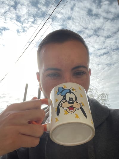 A person is taking a selfie outdoors, holding a mug with a cartoon character on it. The person's face is partially obscured by the mug, but their eyes are visible. They are wearing a gray hooded sweatshirt. The background shows a bright sky with clouds and some power lines. There is a hint of foliage from a tree in the upper right corner. The location is identified as Popintsi, Bulgaria.