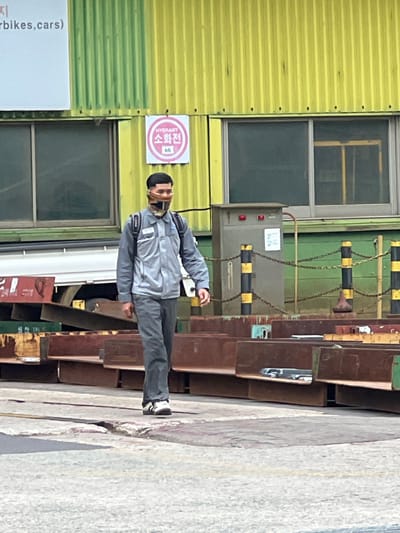 A man wearing a grey work uniform, a patterned mask, and a backpack walks along a paved area in front of a yellow corrugated metal building in Geoje-si, South Korea. The building has large windows and a pink circular sign with Korean text and the number 66 on it. In the foreground, there are stacks of reddish-brown metal beams. A chain-link fence with yellow and black posts separates the man from a utility box attached to the building.