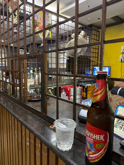 The image shows a bar counter with a Kingfisher beer bottle in the foreground, a clear plastic cup filled with ice and water, and a bottle cap on the counter. Behind the counter, there are several bottles of alcohol, beer crates, and a computer monitor. A metal grate separates the customer area from the bar. In the background, there is a poster or painting of a deity. The scene appears to be in a bar or liquor store. The location is identified as Shamshabad, India.