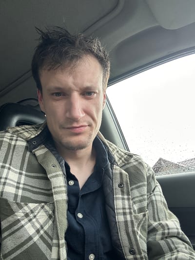 The image is a close-up, slightly tilted selfie of a man sitting in a car. He has short, dark, slightly messy hair and a light beard. He is wearing a dark blue shirt under a green and white plaid jacket with snap buttons. The man is looking directly at the camera with a neutral expression, and his mouth is slightly open. The interior of the car is visible, including part of a sun visor on the left and a dark seat in the foreground. Through the car window behind him, raindrops are visible on the glass, suggesting it is raining. The sky appears overcast and bright. The image was taken in Makiv, Ukraine.
