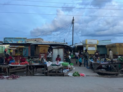 The image depicts a market scene in Kapiri Mposhi, Zambia. Numerous people are present, some of whom appear to be vendors seated behind tables displaying various goods, including tomatoes, onions, dried fish, and leafy vegetables. Customers are visible walking through the market area. The market stalls are constructed from wood and corrugated metal. A sign for "Fair Havens Stationary Shop" is visible above one stall. Several utility poles and power lines are present overhead. The sky is overcast with heavy clouds. Yellow-painted shops with the brand name "Dial" are also seen in the background. The road is paved and appears somewhat worn.