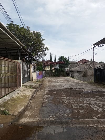 A narrow, cracked road stretches between houses with gated yards. A weathered metal awning and fence stand on the left, while a similar structure is visible on the right. Laundry hangs in the distance near a vibrant purple wall. A large tree casts shade. The sky is overcast, and power lines crisscross overhead, adding to the residential feel.