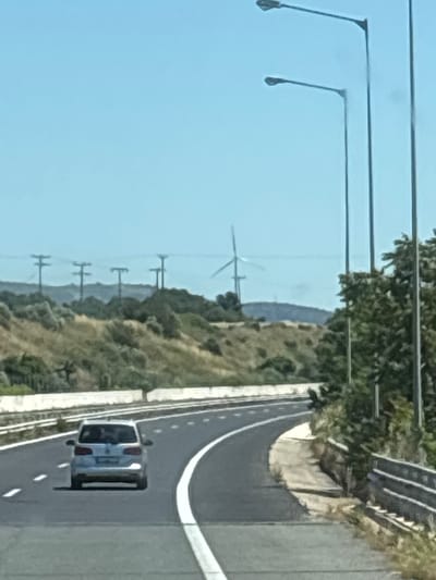 The image depicts a view from a moving vehicle on a highway in Alexandroupolis, Greece. A silver Volkswagen car is driving ahead on the asphalt road, which is marked with white lines. The highway curves to the right, with a white concrete barrier on the right side of the road. Beyond the barrier, there are trees and vegetation. In the background, there are hills with sparse vegetation and several wind turbines are visible. Power lines with supporting poles are also present on the horizon. The sky is clear and blue. The scene suggests a sunny day in a semi-rural area near the city.