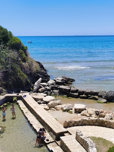The image depicts a scenic view of the sea and a pool area in Sperlonga, Italy. The sea is a vibrant blue, with a small white boat visible in the distance. Rocky shores and lush green vegetation line the coast, creating a natural and picturesque setting.

In the foreground, a shallow pool is bordered by stone structures and worn paving stones. Several people are present in the pool and around its edges. In the water, three young women are visible. One woman stands in the pool, wearing a light-colored swimsuit. Two other women are closer to the foreground, one wearing a dark bikini and the other wearing black shorts and a dark top, with their backs to the camera, appearing to interact with each other.

On the stone pathway bordering the pool, a pair of white flip-flops and a dark blue towel are placed. Another dark bag rests near the edge of the pool. The pool area itself is constructed with a combination of stone and concrete, suggesting it may be a historical or man-made water feature integrated into the natural landscape. The overall impression is a tranquil, sunny day at a coastal location in Sperlonga.