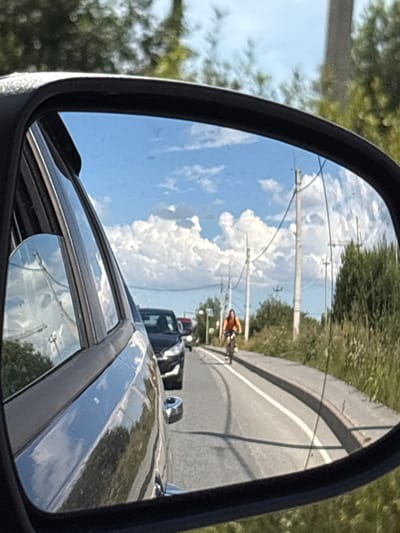 The image shows a reflection in a car's side mirror, depicting a road with a cyclist and other vehicles. The scene appears to be a suburban or rural area, with trees, utility poles, and a cloudy sky. The cyclist, wearing an orange top, is riding on the right side of the road. Other cars are visible in the distance, suggesting traffic. The mirror itself reflects the sky, clouds, and some greenery. The car the mirror is attached to is likely in motion, as evidenced by the blurred background and the reflection. Based on the provided context, this scene is located in the city of Imeni Sverdlova, Russia.
