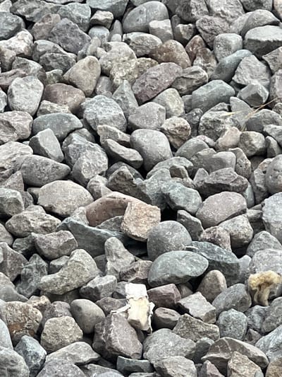 The image displays a dense, textured background of grey and brown river stones, varying in size and shape from small pebbles to larger, irregular rocks. The stones are tightly packed, creating a rugged and natural surface. Some stones have smoother, rounded edges, while others are rough and angular. The lighting appears to be natural daylight, casting subtle shadows that accentuate the three-dimensional quality of the rock pile. There are no visible people or distinct objects other than the stones themselves, suggesting this is a natural or constructed landscape feature. A few stray strands of dry grass or plant material are interspersed among the rocks, adding a touch of organic detail. The overall impression is one of a dry, outdoor environment, possibly a garden path, embankment, or landscaping feature. The location context of Tokmok, Kyrgyzstan, implies this scene is likely found in a region characterized by such geological formations. There is no visible text in the image.