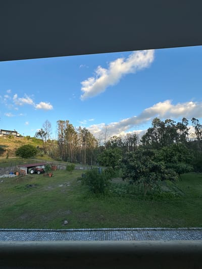 The image shows a view from a window in Celorico de Basto, Portugal. The sky is blue with scattered clouds. A line of trees separates the sky from a green landscape. The landscape includes a grassy area, various trees, and a building on a hillside. Farm equipment is visible in the landscape. The window frame is visible on both sides of the image.