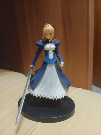 A figurine of Saber from the Fate series is depicted in a dynamic pose, holding a sword. The figurine is predominantly blue and white, with blonde hair and green eyes. It stands on a black circular base, which is placed on a light yellow, textured surface. Behind the figurine, a wooden surface with a horizontal grain pattern serves as the background. The lighting suggests an indoor setting, likely during the day, with soft illumination. The figurine is positioned at an angle, with the sword extending towards the right of the frame. The overall impression is that of a collectible item displayed on a surface.