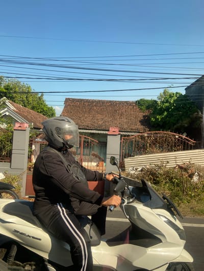 A person wearing a black outfit, a grey helmet with a clear visor, and a black chest bag is riding a white scooter. The person is captured in profile, looking to the right. The scooter's front fairing is white with black accents. In the background, there are residential buildings with terracotta tile roofs and a metal fence. Green trees and foliage are visible, along with a clear blue sky and overhead electrical wires. The scene appears to be a street in a residential area, possibly Bangsal, Indonesia.