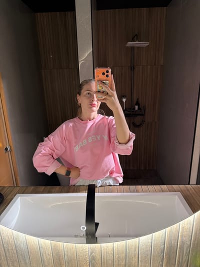 A young woman is taking a mirror selfie in a bathroom. She is wearing a pink sweatshirt and light-colored shorts. Her hair is pulled back in a ponytail. She has on makeup, including red lipstick. She is wearing a watch on her left wrist and a ring on her right hand. The bathroom has wooden walls and a modern sink with a black faucet. The scene is located in a bathroom in the city of Semiluki, Russia.