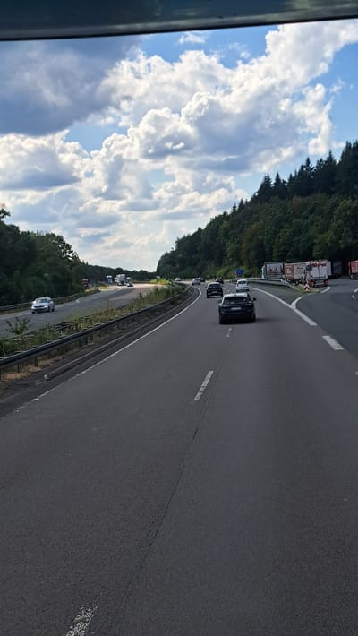 A high-angle shot captures a view from a vehicle traveling on a multi-lane highway in Siegen, Germany. The road is paved and marked with white lines, indicating multiple lanes of traffic. Several vehicles, including cars and trucks, are visible on the highway, moving in the same direction. The highway is bordered by a metal guardrail on the left and a dense, green forest on the right, with a cloudy sky overhead. The scene depicts a typical highway commute or travel scenario.