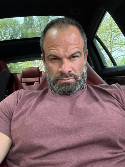 A man with a graying beard and thinning hair is in a car with a sunroof open. He is wearing a maroon t-shirt and looks towards the camera. The car has red leather seats, and trees are visible through the sunroof. The location is not discernible as being in the city of Diekirch, Luxembourg, based on the image.