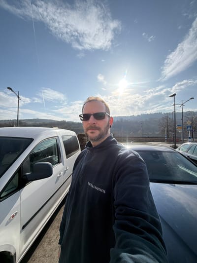 An adult male is prominently featured in a daytime outdoor setting, likely a parking area in Grevenmacher, Luxembourg. He is positioned centrally, facing the camera, suggesting a selfie. The man has a beard, short hair, and wears dark-tinted sunglasses and an earring in his visible left ear. He is dressed in a dark blue long-sleeved quarter-zip pullover, displaying "PONTS & CHAUSSÉES" (Roads and Bridges) in white text on the left chest.
To his immediate left, a white passenger van or minivan is partially visible. To his right, the hood and windshield of a dark-colored car are present. The background reveals a bright blue sky with scattered white clouds and multiple aircraft contrails. A radiant sun, positioned high, creates a lens flare effect. Beyond the vehicles, a landscape of tree-covered hills rises, characteristic of the Moselle valley. Two streetlights with multiple arms are visible, along with glimpses of other vehicles and partial road signs, indicating a public thoroughfare or parking facility. The environment is consistent with a sunny day in a European town, specifically Grevenmacher due to the "Ponts & Chaussées" uniform and geographical features.