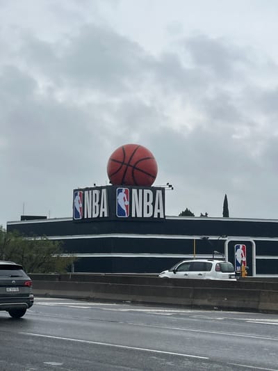 The image depicts the NBA Argentina headquarters in Martínez, Argentina. The large NBA sign with a basketball on top is visible against a cloudy sky. A highway runs alongside the building, with cars in motion. The building's facade is gray and black.
