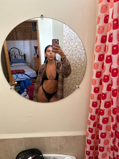 The image shows a woman standing in a bathroom, taking a selfie in a circular mirror. She is wearing a black bikini top and bottom and a plaid button-down shirt. The bathroom has tiled walls and a shower curtain with a pattern of red teddy bears. The location of the bathroom is Montañita, Ecuador, with the coordinates -1.825 latitude and -80.753 longitude. A bed with a black metal frame is visible in the mirror, along with some luggage and other items. The woman's left hand is holding a smartphone, while her right hand is on her hip. She is looking at the camera and appears to be smiling. The mirror reflects the bathroom with a tiled wall behind her.