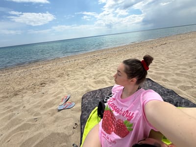 A woman is sitting on a beach in Νέα Φλογητά, Greece. She is wearing a pink t-shirt with a raspberry print and has her hair up in a bun with a red scrunchie. She is sitting on a dark gray blanket with a lime green towel underneath. A pair of striped flip-flops are next to her on the sand. The beach stretches out to the left, with the sea and sky in the background. The sky is partly cloudy.