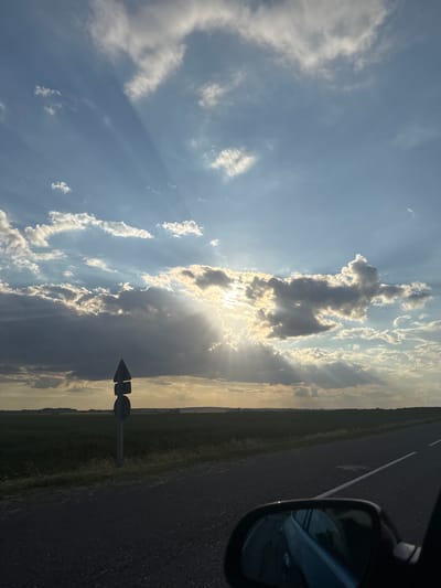 The image depicts a roadside scene in Belarus, likely during late afternoon or early evening, as indicated by the warm, diffused sunlight breaking through the clouds. The sky is a prominent feature, with a mix of bright white and dark grey clouds creating dramatic contrasts. Sun rays are visible, fanning out from behind a dense cloud formation.
The scene includes a paved road with a white dividing line, suggesting a highway or a significant road. To the right of the road, a car's side mirror is visible, reflecting the sky and surrounding environment. The area adjacent to the road is a field of green grass and vegetation, extending towards the horizon. A solitary signpost, shaped like an arrow, is situated in the grassy area.
The overall mood is serene and contemplative, possibly evoking a sense of journey or travel. There is no visible text in the image. The time of day is suggested by the quality of light, with the sun low on the horizon. The weather appears to be partly cloudy. The location is identified as Азгінавіцкі сельскі Савет, Belarus.