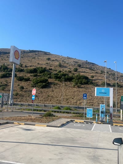 The image shows a street view in the Municipal Unit of Amphilohia, Greece, on a clear, sunny day with a bright blue sky. In the foreground, there is a concrete forecourt with yellow and gray curbs, and tire marks are visible on the concrete. To the right, the side mirror of a silver car is partially visible.
Further back, there is a dedicated electric vehicle charging station with two blue charging units and accompanying signage that reads "incharge." One sign also indicates "Electric vehicle charging." Next to the charging station, there is a fire extinguisher mounted on a pole. There are also parking signs, including a blue sign with a white "P" and a red and white octagonal "STOP" sign with an arrow pointing right below it.
To the left of the charging station and signs, a tall pole with a Shell logo is prominent. Behind these elements, a highway overpass with metal guardrails is visible, separating the charging area from a dry, grassy hillside covered with scattered bushes and trees. The hillside appears rocky and sparsely vegetated. Streetlights are positioned along the road and near the charging station.