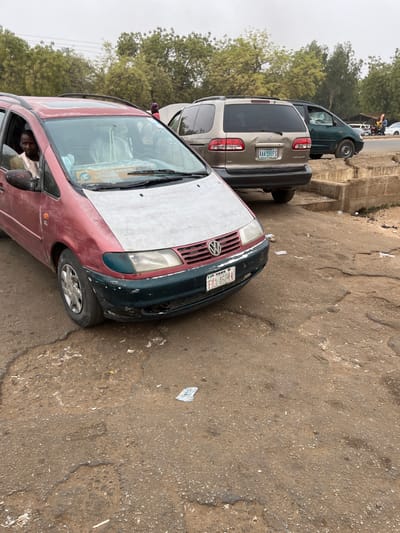 The image depicts an outdoor scene in Kasuwan Kaji, Nigeria, featuring multiple parked minivans on a degraded, unpaved or heavily potholed asphalt surface.
In the foreground, slightly angled towards the right, is a maroon (dark red) Volkswagen Sharan minivan. Its hood is a contrasting faded light grey or unpainted, and the front bumper is dark green, showing extensive scuffing, chipping, and damage, including a missing section on the lower passenger side. The vehicle bears a front license plate "FKA 460 KA" with "TFP YOB" above it. A male individual with a beard, wearing a light-colored garment, occupies the driver's seat, looking directly towards the camera. Indistinct figures are visible in the rear passenger seats. A package, possibly labeled "Peace Essential," rests on the dashboard. The driver's side front tire appears underinflated.
Behind and to the right of the Volkswagen is a gold/tan Toyota Sienna minivan, parked facing away from the viewer. Its rear bumper exhibits a noticeable dent on the passenger side. This vehicle's license plate reads "AAA-591FS" with "ABUJA" displayed above. Partially visible between these two vehicles, near the front of the Toyota Sienna, is a person wearing a red head covering.
Further in the background, to the right of the Toyota Sienna, a dark green minivan is partially visible, parked on what appears to be a slightly elevated, cleaner road surface. A low, damaged concrete barrier or curb separates the foreground area from this elevated section.
The ground surface is uneven, dark brown/grey, and littered with various small fragments of white paper or plastic. The background features numerous green trees with sparse foliage under an overcast sky.
