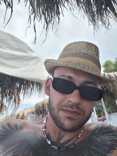 A man wearing a straw hat and sunglasses is captured in a close-up, selfie-style shot. He has tattoos on his chest and shoulders and is wearing a silver chain. The background suggests an outdoor, possibly beachside, location with palm fronds and a white umbrella providing shade. The sky is overcast. The man is positioned centrally in the frame, looking towards the camera. The image appears to have been taken in the Municipal Unit of Diakopto, Greece, given the distinctive vegetation and architectural style of the beach umbrellas.