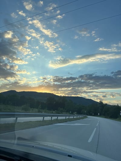 The image shows a road curving through a rural landscape. The road has white lane markings and a metal guardrail along the side. In the background, there are rolling hills covered in trees, and a sunset is visible in the sky. The sky is a mix of blue with scattered clouds, some of which are illuminated by the setting sun. There are power lines visible in the upper portion of the image. The scene appears to be a view from a vehicle traveling on the road. The specific location in the city of Kaleytsa, Bulgaria is not identifiable from the visual information provided. There are no people or specific actions depicted in the image.