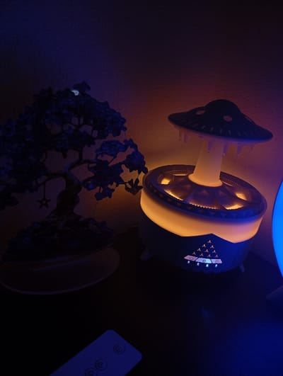 A black bonsai tree sits on a white base, with a decorative, white, mushroom-shaped lamp with a glowing base next to it on a dark wooden surface. The lamp has white geometric designs on its base and a glowing, light-orange inner ring. The scene is lit with a soft, blue light, indicating a night setting. There is a remote control on the bottom of the image. The location is a private residence in Dunajská Lužná, Slovakia, based on the provided coordinates.