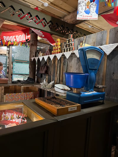 The image depicts a section of a museum exhibit, likely simulating a Soviet-era shop or market stall. The scene is set indoors, with a rustic wooden-planked wall serving as a backdrop.

Dominating the right side of the frame is a vintage blue mechanical scale, with a weighing pan and a dial indicator. In front of it, a blue basin is placed on the scale. To the left of the scale, on a wooden shelf, are stacks of canned goods and a small framed picture. Further to the left, hanging on the wall, are various bags and dried items.

In the foreground, a glass display case contains what appears to be red fabric with white lettering, possibly an advertisement or product display. Above this, on a wooden counter, is a wooden abacus with numerous beads.

The ceiling area reveals decorative wooden elements and banners, one red banner with Cyrillic lettering is prominent. A poster featuring two young individuals is also visible near the ceiling. A woman is partially visible on the far left, looking towards the exhibit. The overall atmosphere suggests a historical reenactment or display of everyday life from a past era. The location is identified as Ust'-Kabyrza, Russia.