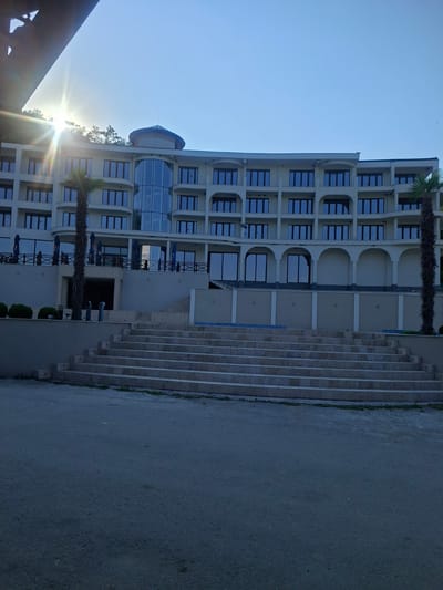 The image depicts a large, modern hotel building with a light-colored facade and multiple floors, featuring numerous windows with dark frames. The architecture includes curved balconies and an entrance area with arched openings on the ground level. A prominent feature is a glass-enclosed section on the building's front. In front of the hotel, a wide, multi-tiered staircase made of light-colored stone leads up to the entrance level. Flanking the staircase are palm trees. The area in front of the staircase is paved with asphalt. To the left of the frame, a portion of a dark, angular structure is visible, possibly a roof overhang or a part of a different building. The sky is clear and blue with the sun shining brightly from the upper left, casting a lens flare. The scene appears to be set in Pisanets, Bulgaria, suggested by the location context. No people are visible in the image.
