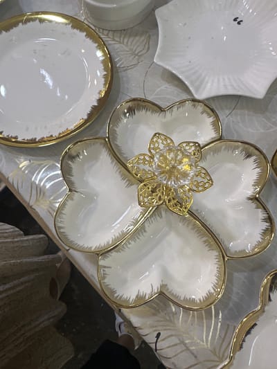 A white ceramic serving dish with a gold trim, shaped like a flower with four heart-shaped compartments, sits on a table. A decorative gold and clear glass flower sits in the center of the dish. In the background, other white ceramic plates and dishes are visible. The table is covered with a patterned cloth with gold leaf designs. The scene is indoors, likely a market or shop, given the presence of multiple items for sale and a person's feet visible in the lower right corner, suggesting a retail environment. The image was taken in Kara-Suu, Kyrgyzstan.