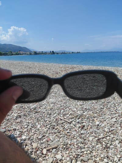 An outdoor, eye-level, close-up shot captures sunglasses being held in the foreground against a blurred backdrop of a sun-drenched beach, a tranquil blue sea, and a distant coastline dotted with buildings and trees under a clear blue sky with scattered white clouds. The sunglasses lenses reflect the pebbled shore.