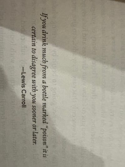 The image displays a close-up, angled view of a page from a book. The page is light-colored, possibly off-white or cream, and features black text printed in a serif font. A slight diagonal shadow is visible across the upper left and central portions of the page, indicating uneven illumination or a slight curvature of the page.
The visible text is a quote, horizontally oriented if the image were rotated ninety degrees clockwise, and reads:
"If you drink much from a bottle marked 'poison' it is
certain to disagree with you sooner or later.
—Lewis Carroll"
No people, specific objects other than the book page itself, or discernible actions or interactions are present in the scene. The background consists of a blurry, light-colored surface, likely another page or the opposing side of the open book. The specific location within the city of McAllen, United States of America, cannot be determined from the provided image.