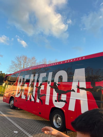 A red Irizar coach bus, emblazoned with the white lettering "BENFICA" and "SPORT LISBOA E BENFICA" on its side, is parked on a paved surface in Leiria, Portugal. The bus's red body features black silhouette decals of athletes engaged in various sports, including football, volleyball, and handball. A partial web address, "BENFICA.PT", is visible on the lower front section. The scene is outdoors under a blue sky with scattered white clouds. Leafless deciduous trees line the background, with some evergreen foliage visible on a rising incline. The ground is a paved parking area marked with white lines. In the lower right foreground, the right hand and the back of a person's head, wearing a dark cap, are partially visible.